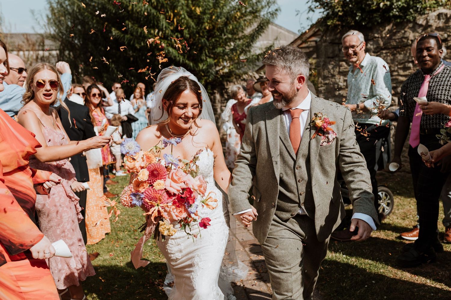 Wedding celebration outdoors on a sunny day with guests throwing flower petals on a happy couple walking at Lyde Court, Herefordshire.
