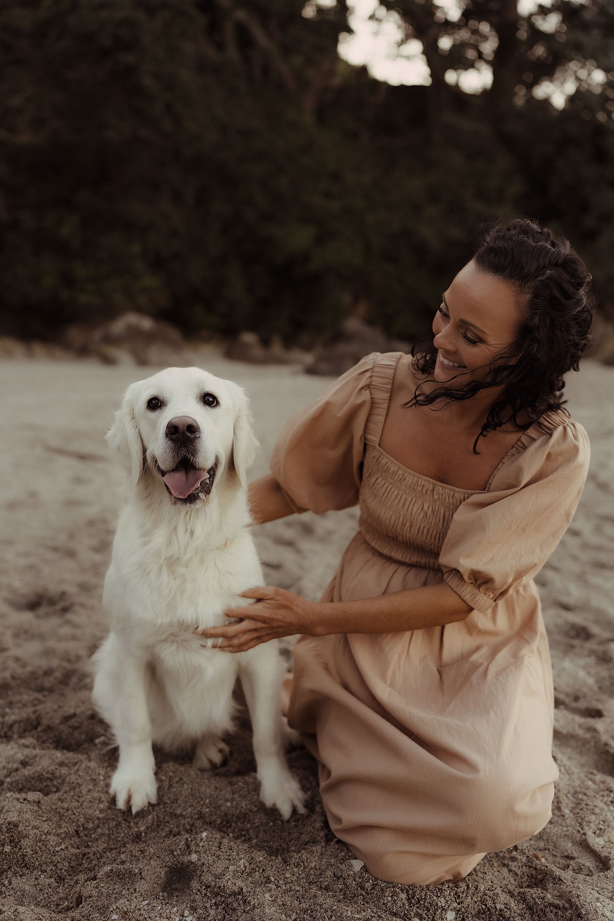 A white labrador retriever sits on a sandy beach with companion wearing a flowing beige dress at sunset.