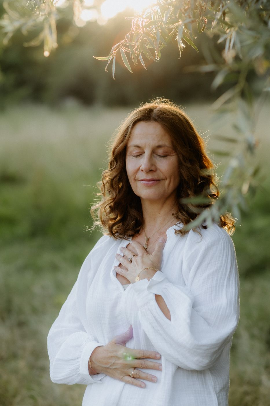 A serene woman with wavy brown hair stands in a sunlit meadow, eyes closed and hands gently placed on her chest and stomach.