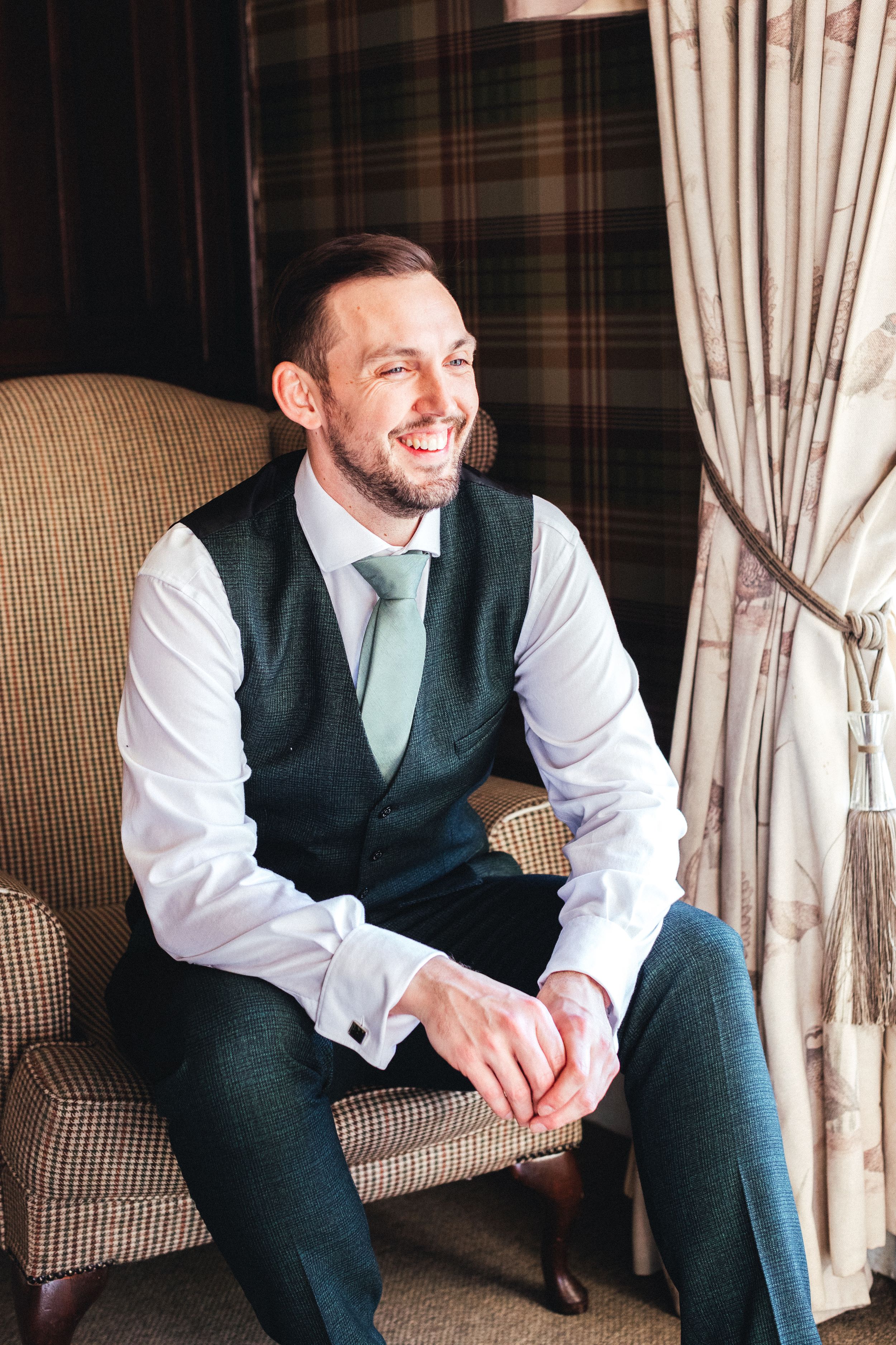Wedding guest in formal attire seated in elegant armchair with plaid curtains.