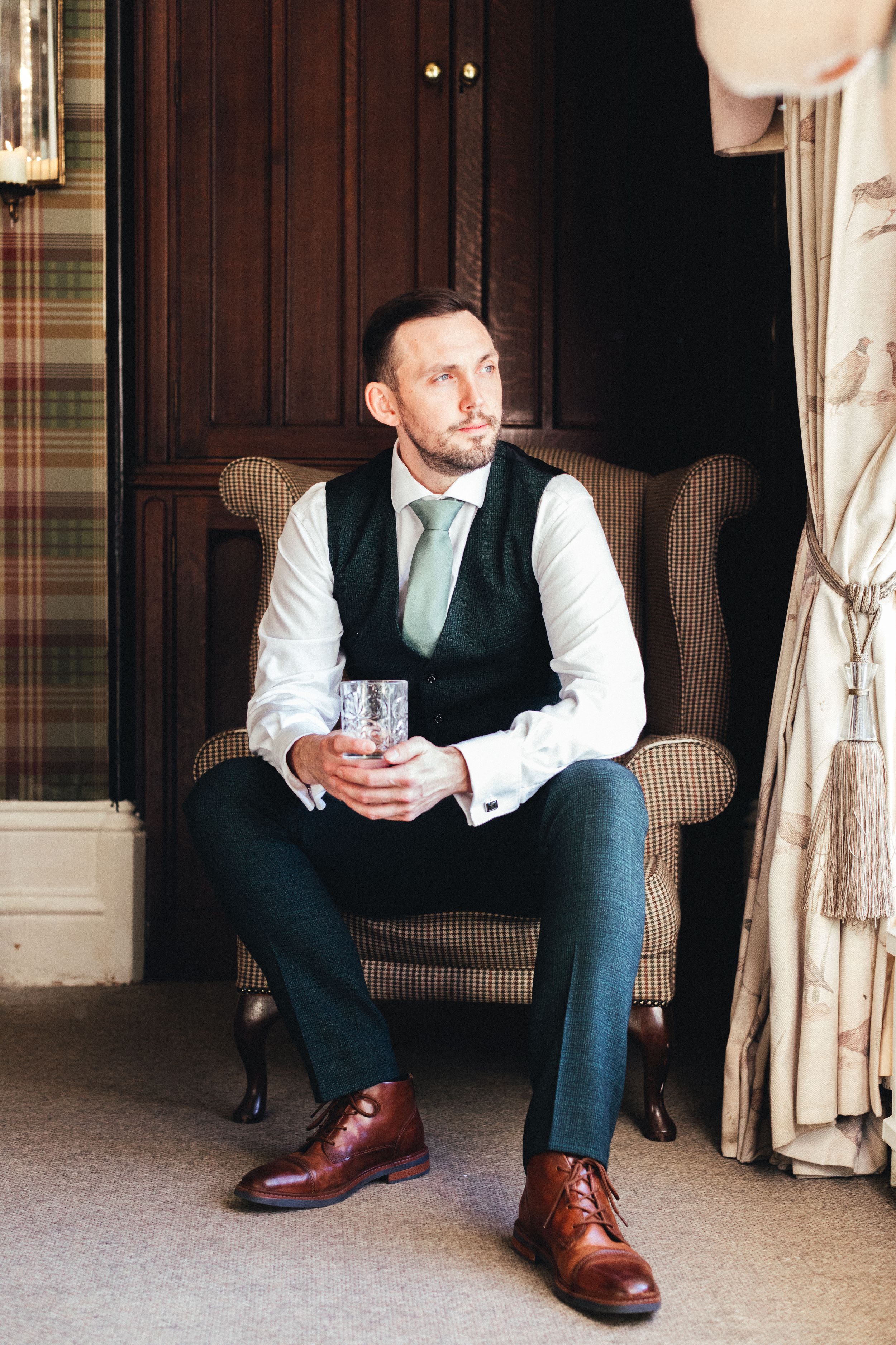 Groom in formal attire relaxes in vintage armchair holding crystal glass.