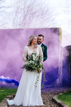 Wedding couple poses amid purple smoke effects in outdoor garden setting.