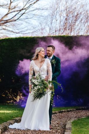 Bride in lace gown and groom in navy suit pose against purple smoke bombs in garden setting during wedding photoshoot.