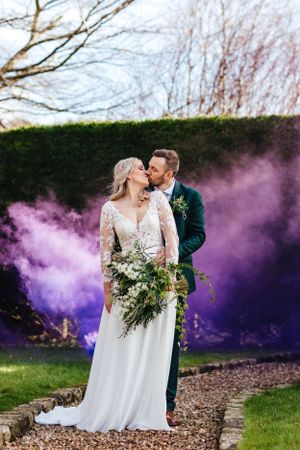 Newlyweds share intimate moment surrounded by dramatic purple smoke in outdoor garden setting.
