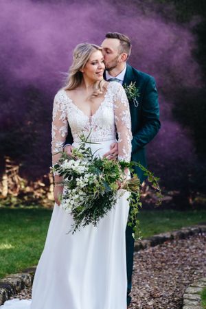 Romantic wedding portrait with purple smoke effect featuring elegant couple and natural greenery bouquet.