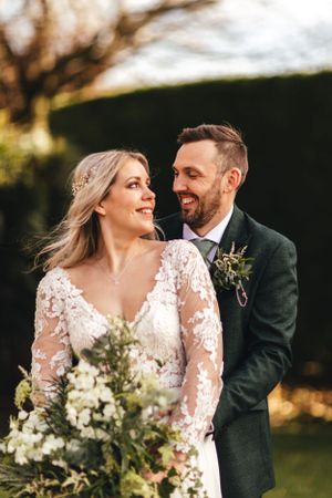 Joyful wedding portrait featuring elegant lace gown and natural floral bouquet against garden backdrop.