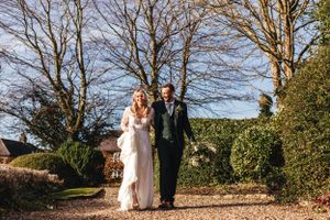 Wedding couple walking along garden path surrounded by bare winter trees and manicured hedges.