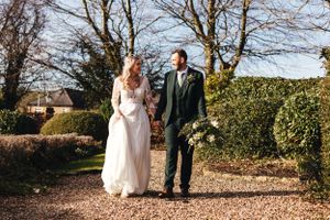 Wedding couple strolling hand in hand down garden gravel path surrounded by winter foliage.