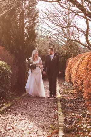 A newlywed couple walks along an autumn garden path lined with orange foliage and bare tree branches.