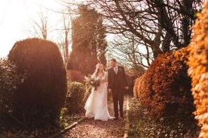 Wedding couple strolls through a scenic garden pathway surrounded by bushes and trees at sunset.