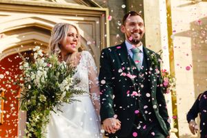 Happy newlyweds exit church while pink confetti showers around them in celebration.
