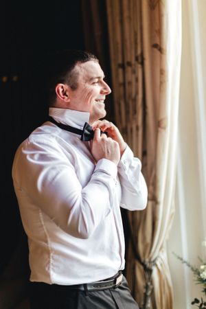 A person adjusts their bowtie while getting ready in a formal white dress shirt by a window.