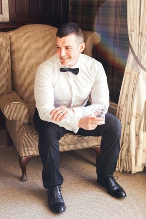 A person in formal attire sits in a plaid armchair holding a glass.