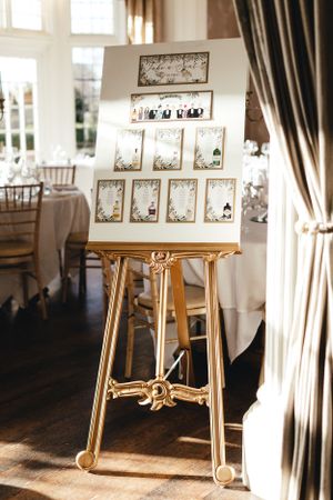 An elegant gold easel displays a wedding seating chart by a window in a reception venue.