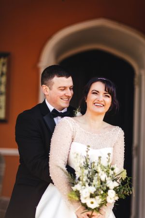 A joyful wedding couple shares a tender moment while holding a white floral bouquet.