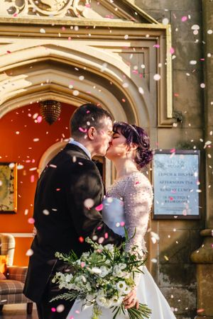 Romantic wedding couple sharing a kiss under gothic archway with falling flower petals and elegant white bouquet.