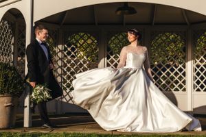 Elegant wedding dress billows in the wind as couple poses in garden gazebo during sunset.