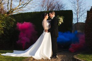 Wedding couple embraces with colorful smoke bombs creating pink and blue effects in garden setting.
