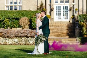 Sunlit wedding portrait with purple smoke effect in front of elegant stone manor.
