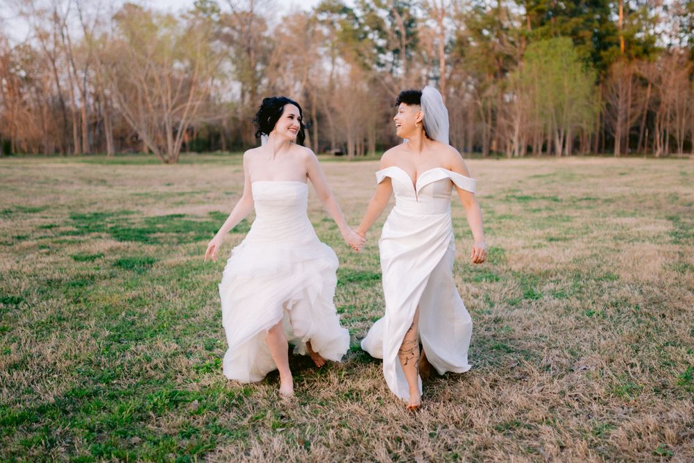 Two elegantly dressed figures run joyfully hand-in-hand through a grassy field on a sunny day.
