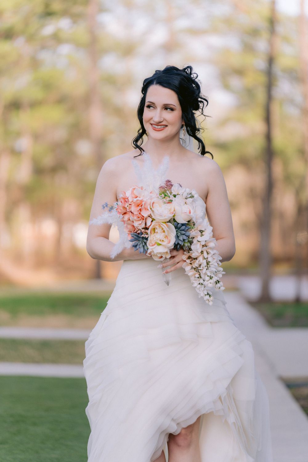 Bride in flowing white dress holds peach and cream bouquet in outdoor setting.