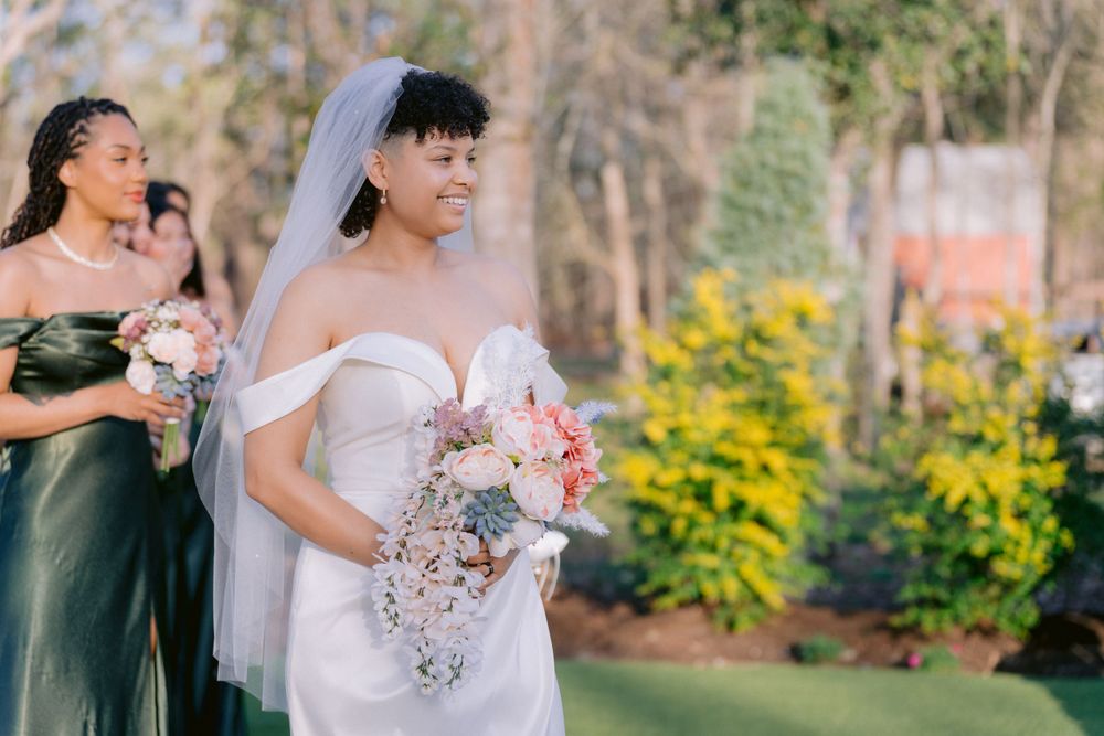 Two people at an outdoor wedding ceremony with one wearing a white gown and holding a pink bouquet.