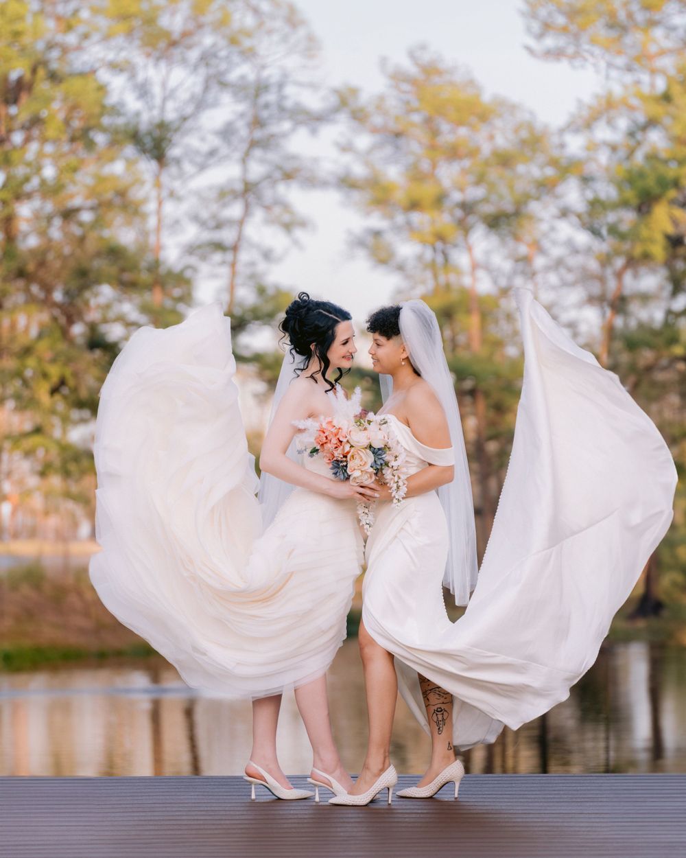 Two brides in flowing white dresses pose on dock with wind-swept fabric.