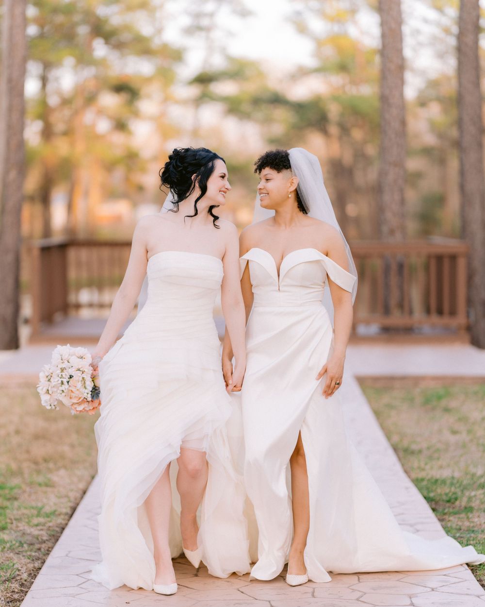 Two brides in elegant white wedding gowns walk hand in hand on wooden walkway.