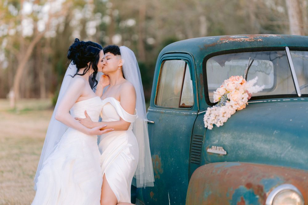 Two brides in white dresses pose by vintage turquoise truck with floral bouquet.