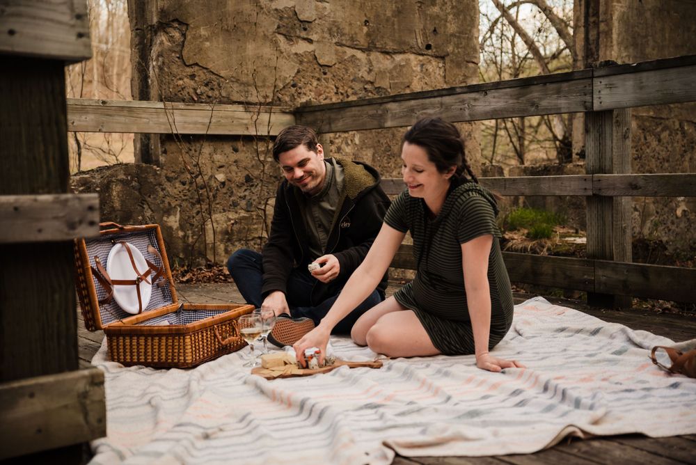 Couple sharing food during cozy picnic on blanket with wicker basket near stone wall.