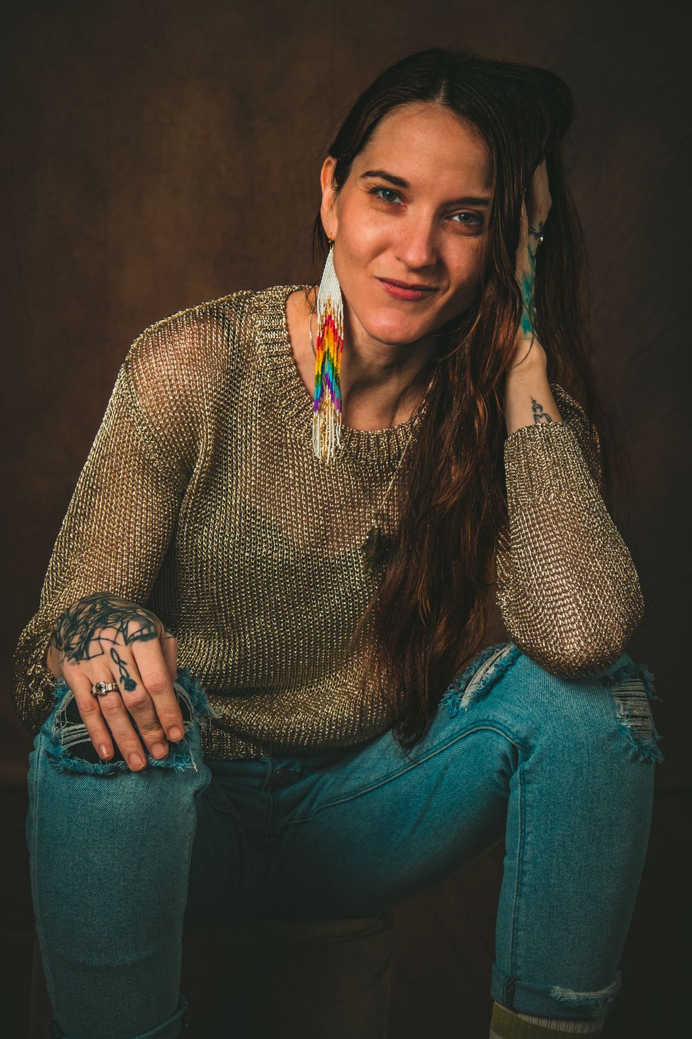 Casual portrait showing colorful feather earrings, knit sweater, and multiple rings in relaxed pose.