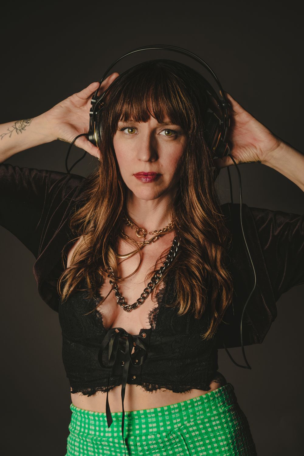 Fashion portrait with headphones, chain necklace, black top and green patterned pants against dark backdrop.