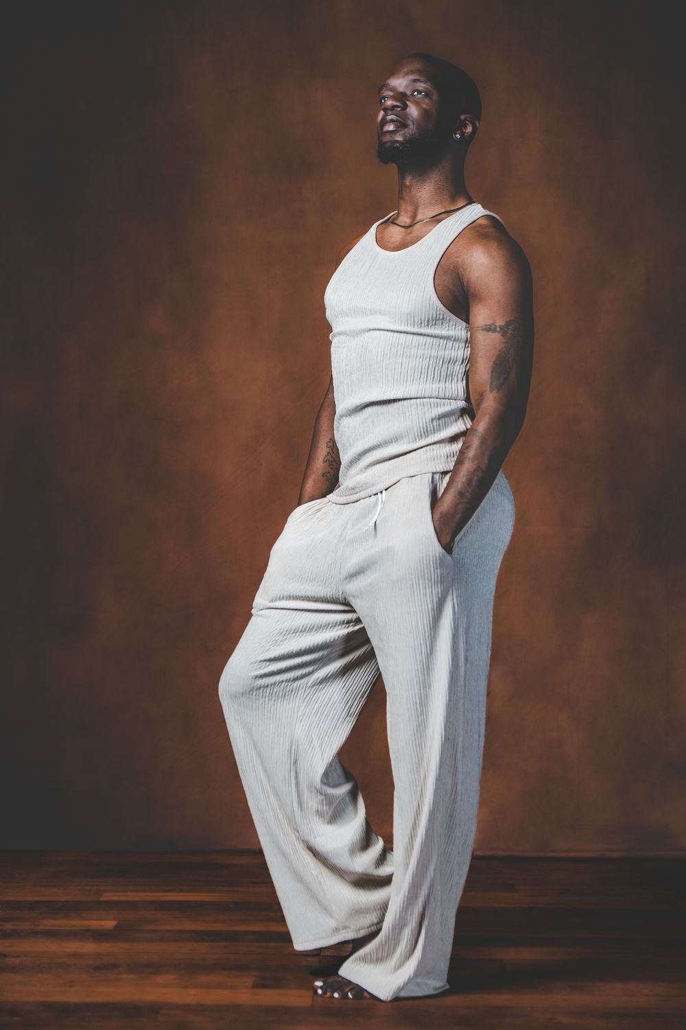 Athletic figure in white tank top and loose pants poses confidently against warm brown backdrop.