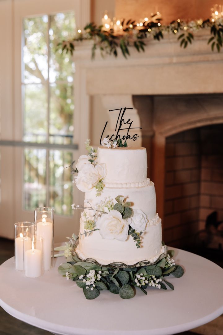 An elegant three-tiered white wedding cake decorated with fresh flowers and greenery sits beside lit candles.