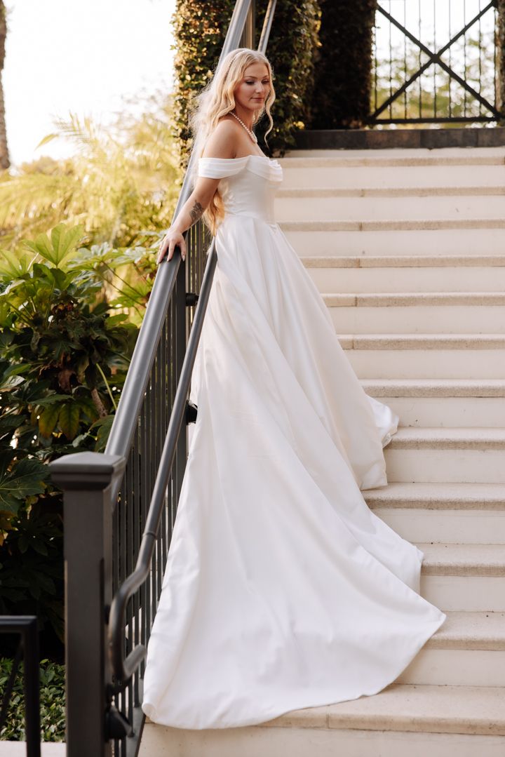 Elegant off-shoulder white wedding gown with flowing train photographed on outdoor stone staircase at golden hour.