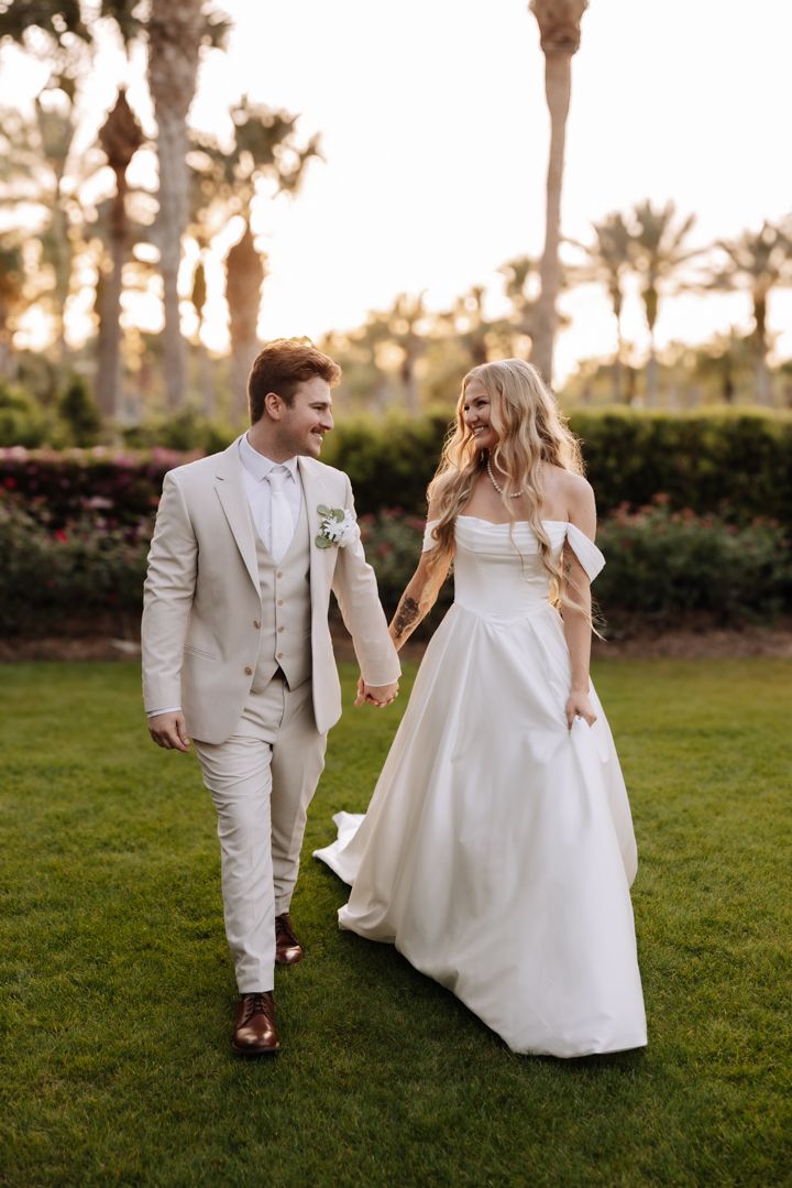 A couple walks hand in hand through a palm-lined garden during golden hour, wearing light-colored wedding attire.