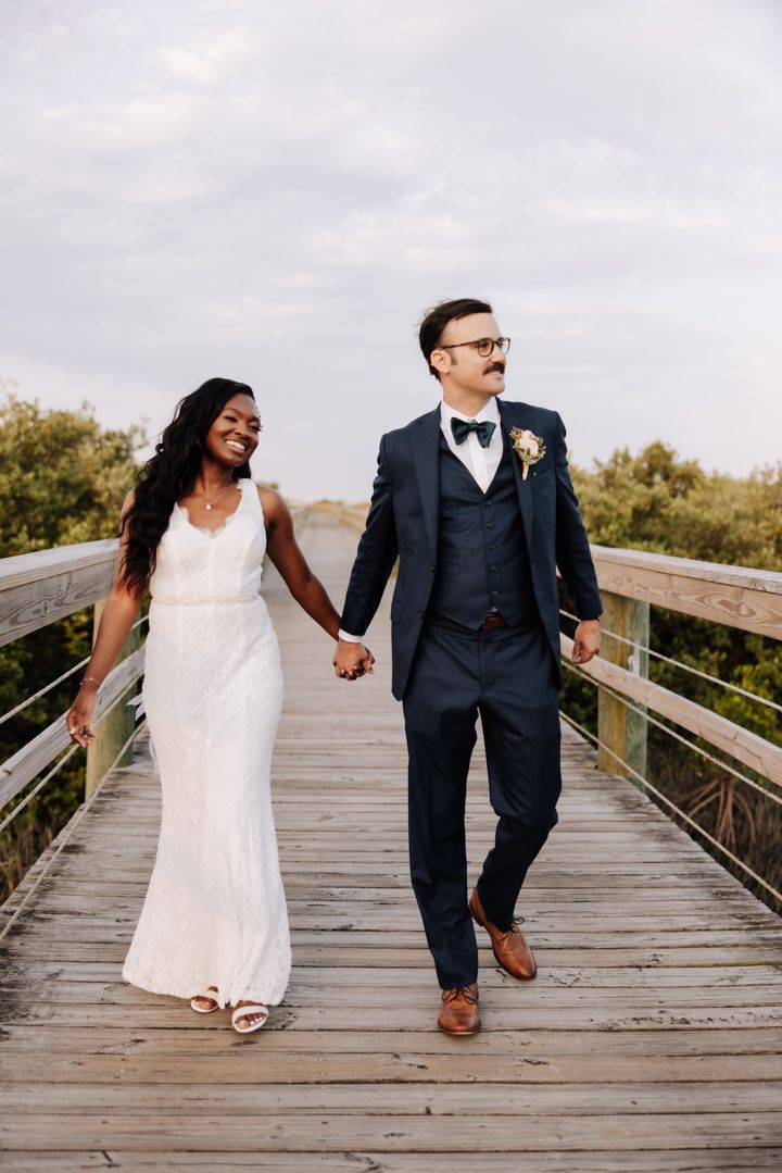 Wedding couple walks hand in hand laughing on a rustic wooden boardwalk during golden hour.