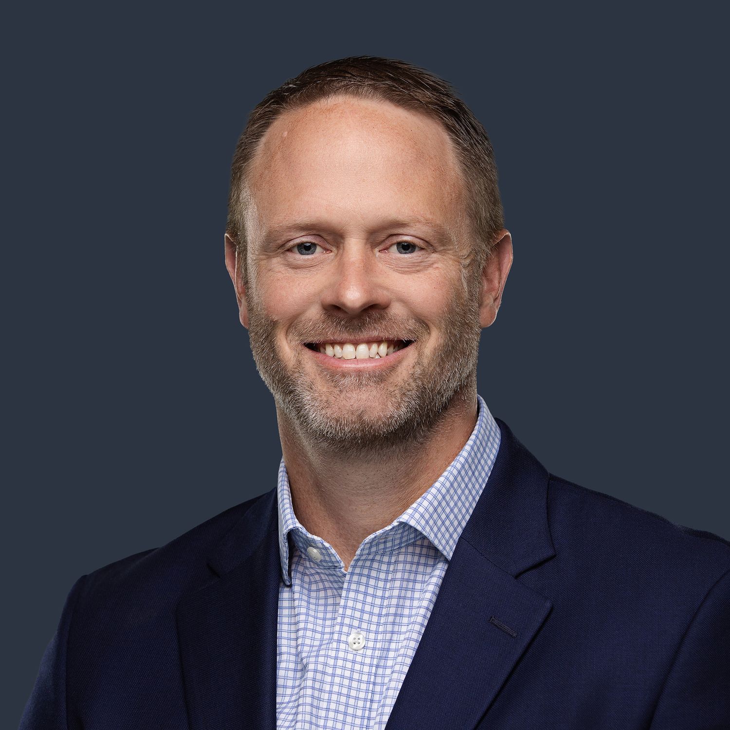 Professional headshot against dark background wearing navy blazer and checkered shirt.