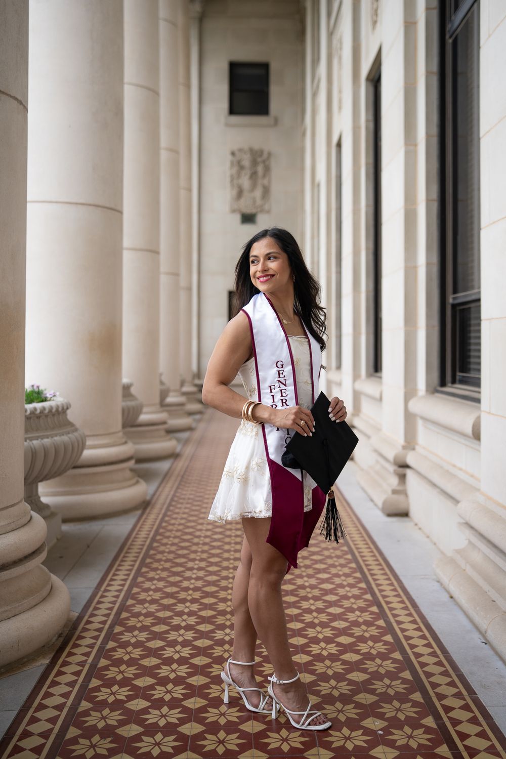 A graduate in a white sash and burgundy dress stands elegantly in a columned hallway with ornate patterned tile flooring.
