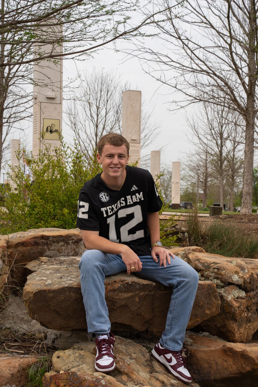 College student in Texas A&M jersey number 12 and jeans sits on large rocks with bare trees in background.