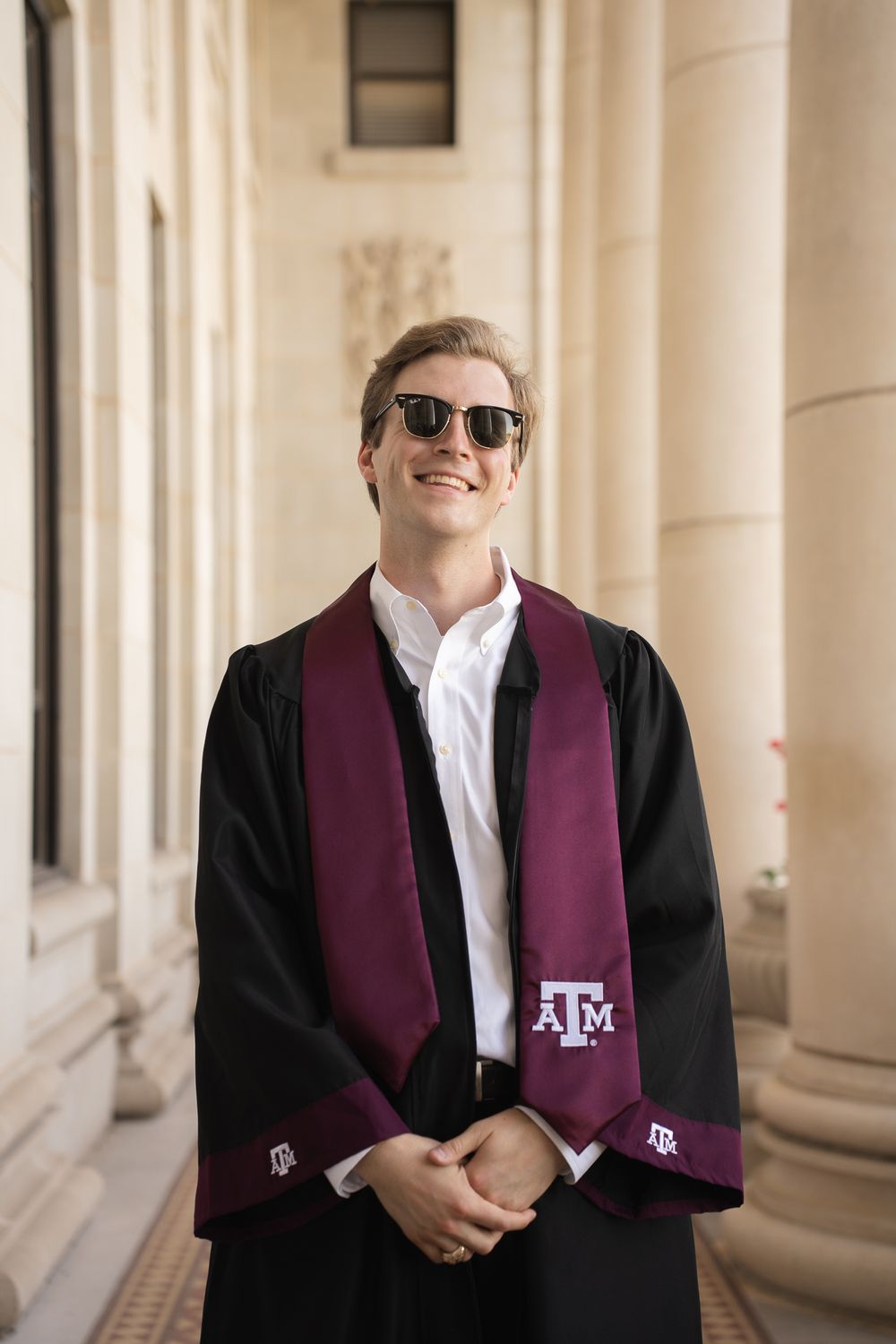 Graduate wearing a Texas A&M graduation stole and black sunglasses smiles while posing between classical columns.