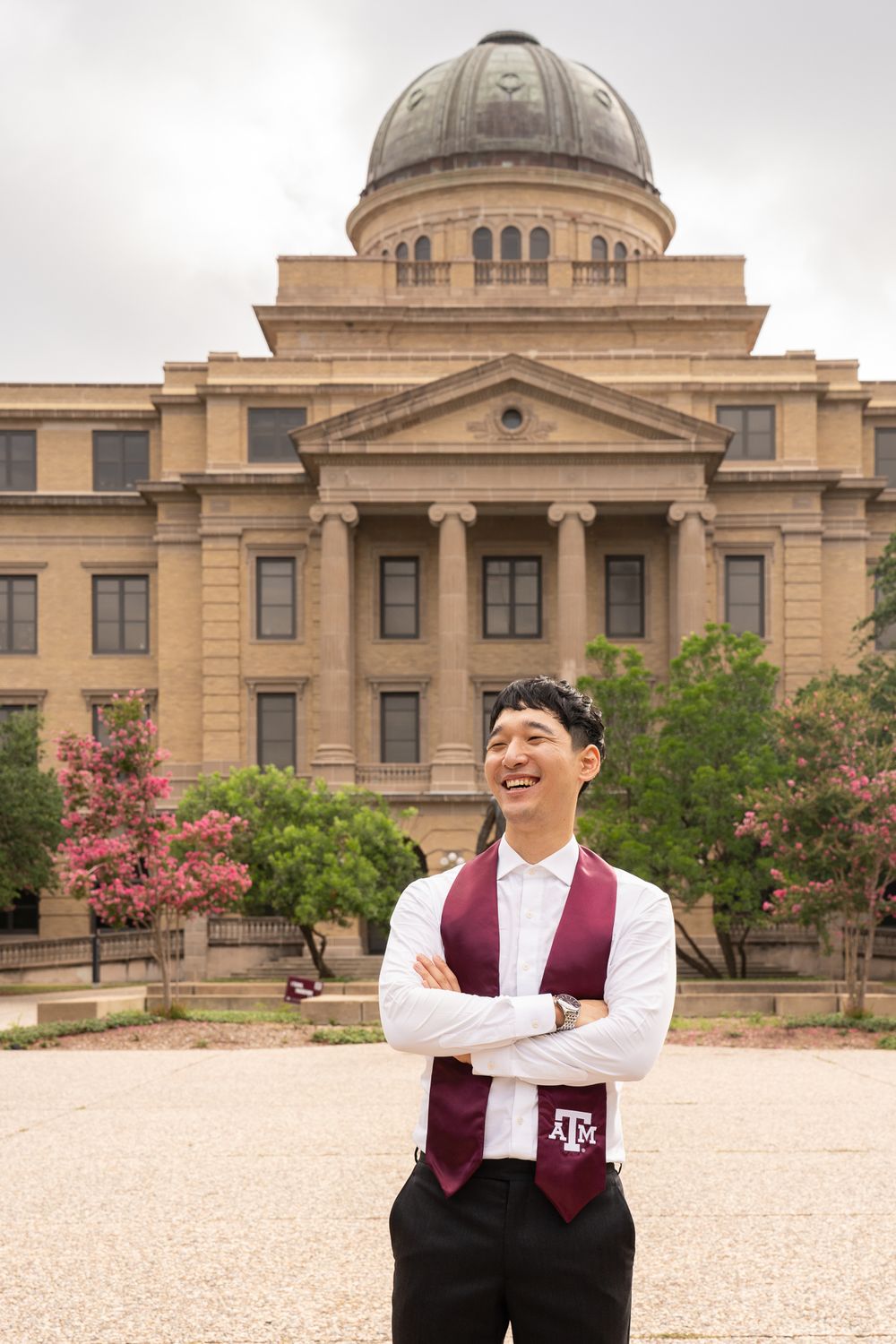 A graduate wearing maroon and white Texas A&M colors poses confidently in front of the Academic Building on campus.