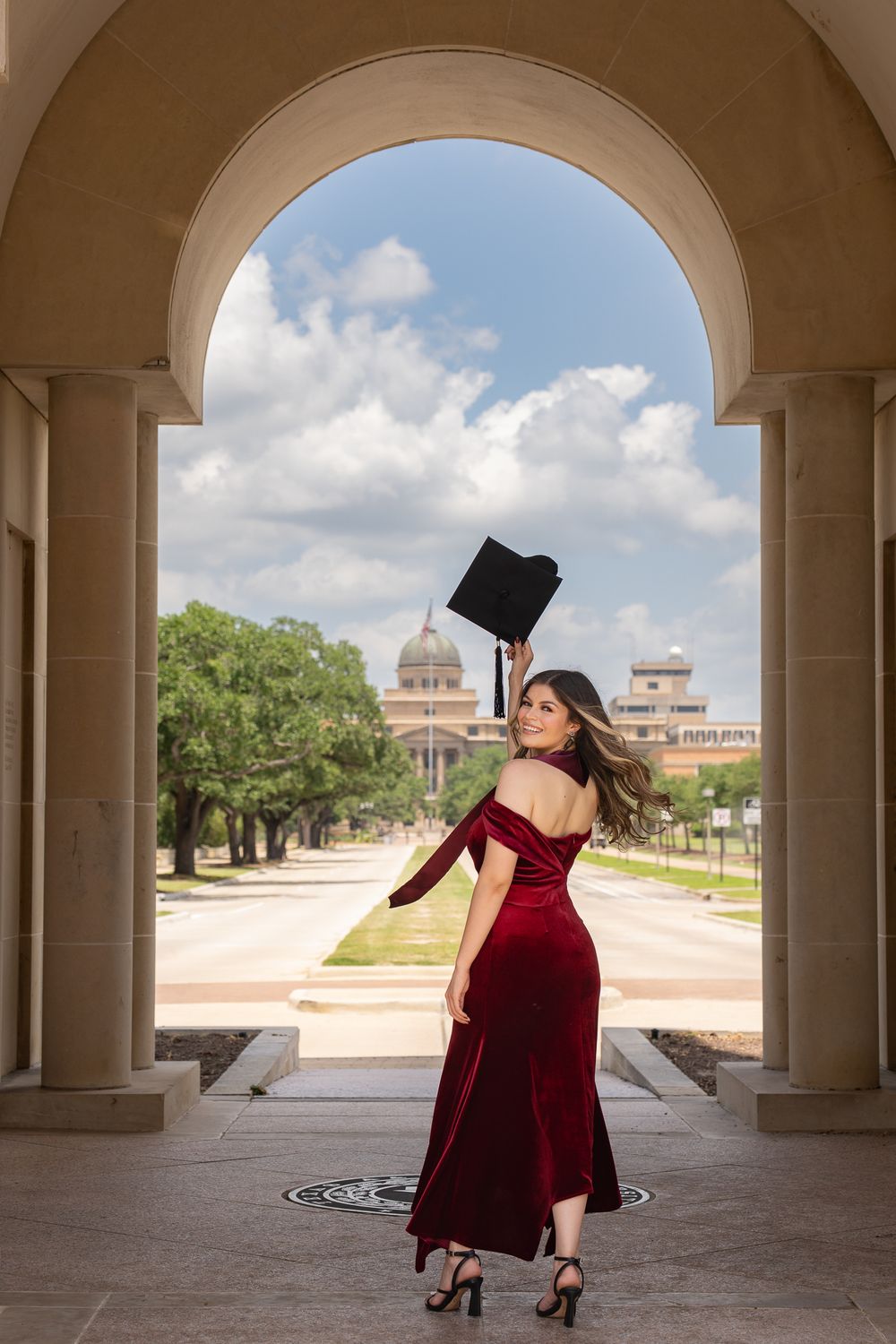 A graduate in a red velvet dress tosses their graduation cap under an arched walkway with classic architecture.