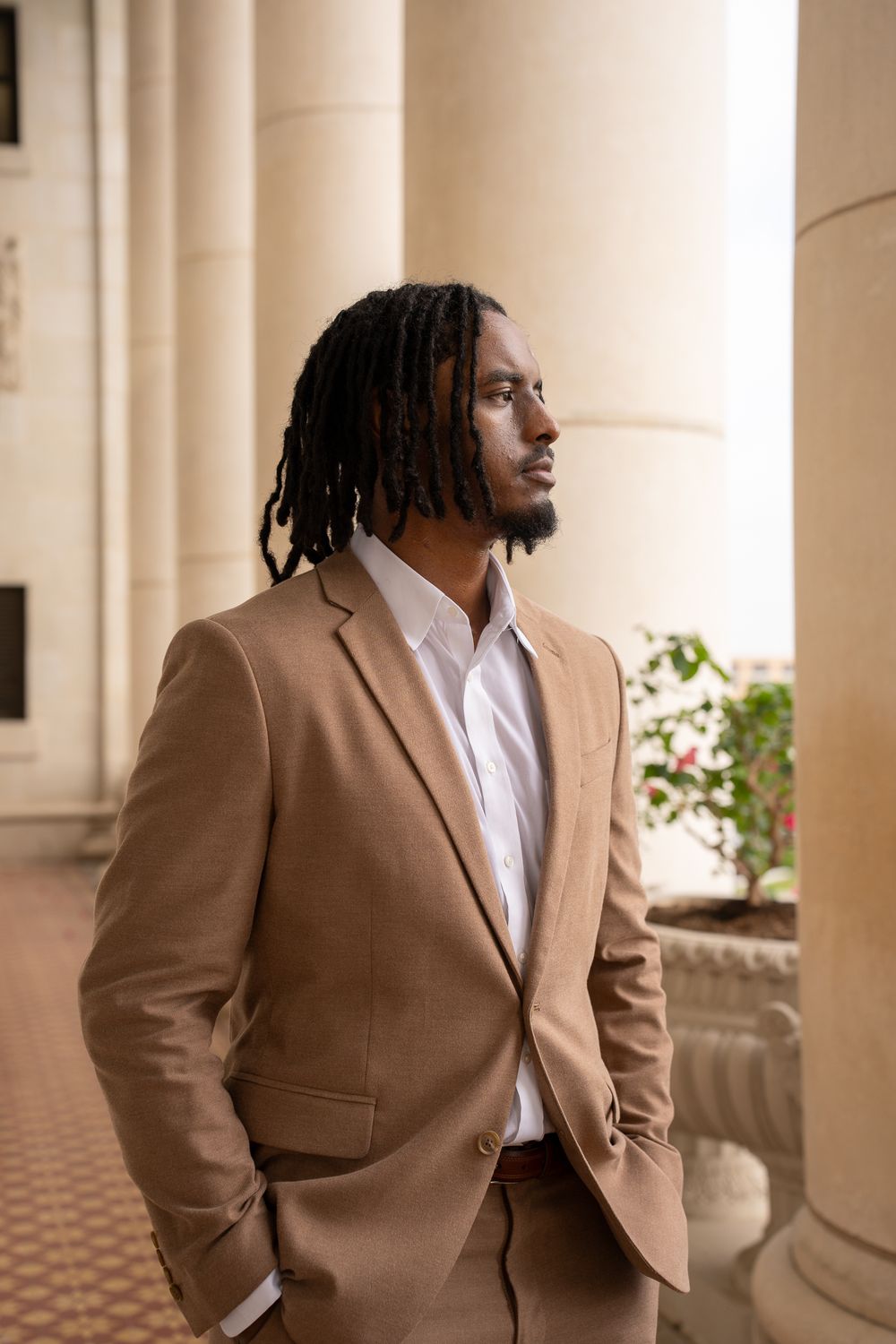 Stylish man wearing tan suit jacket and white dress shirt poses thoughtfully in elegant building with columns.
