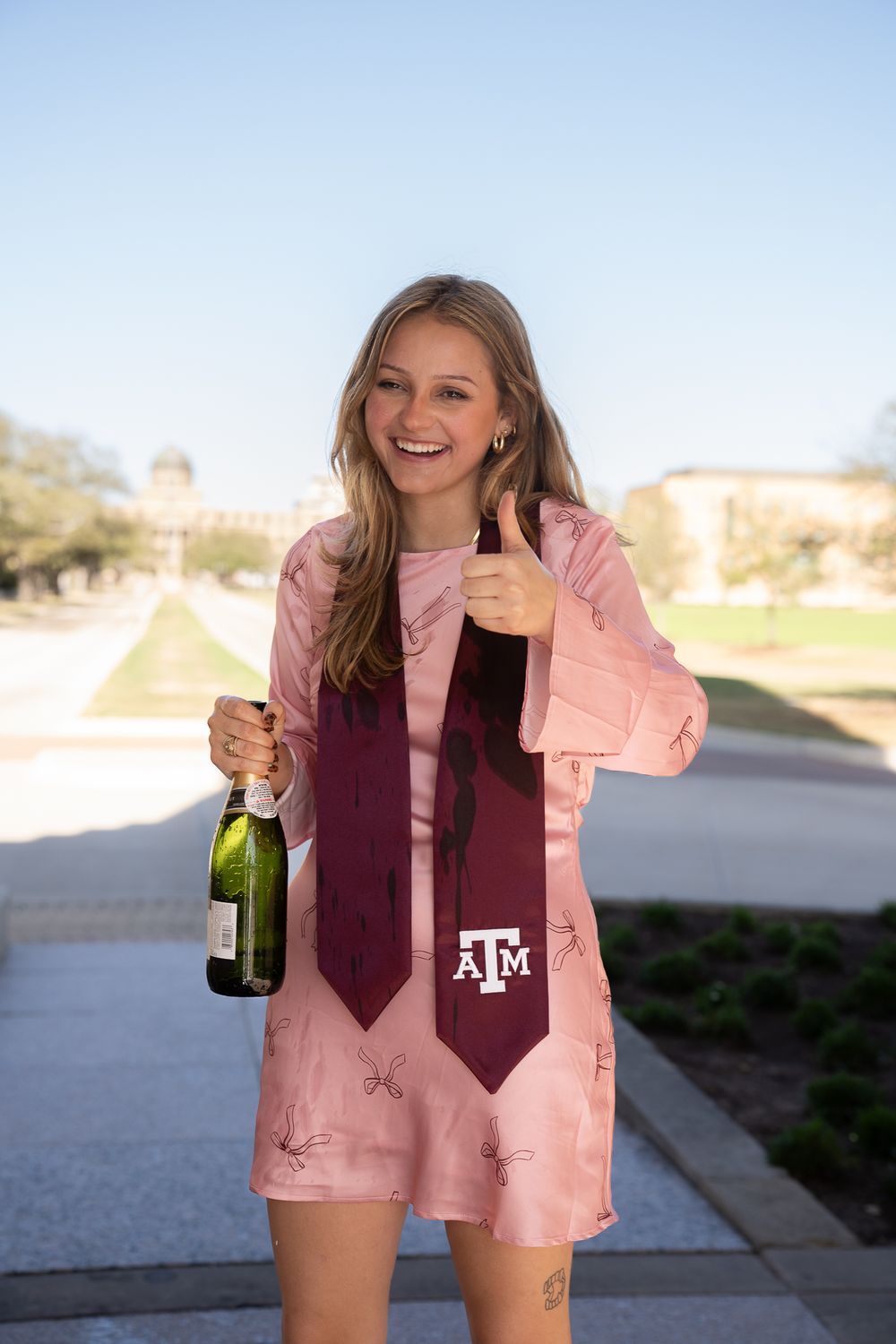 Graduate in pink outfit celebrating with champagne and Texas A&M tie on campus.