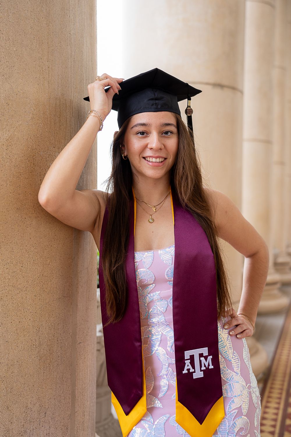 A Texas A&M graduate in cap and gown poses confidently against a stone column backdrop.
