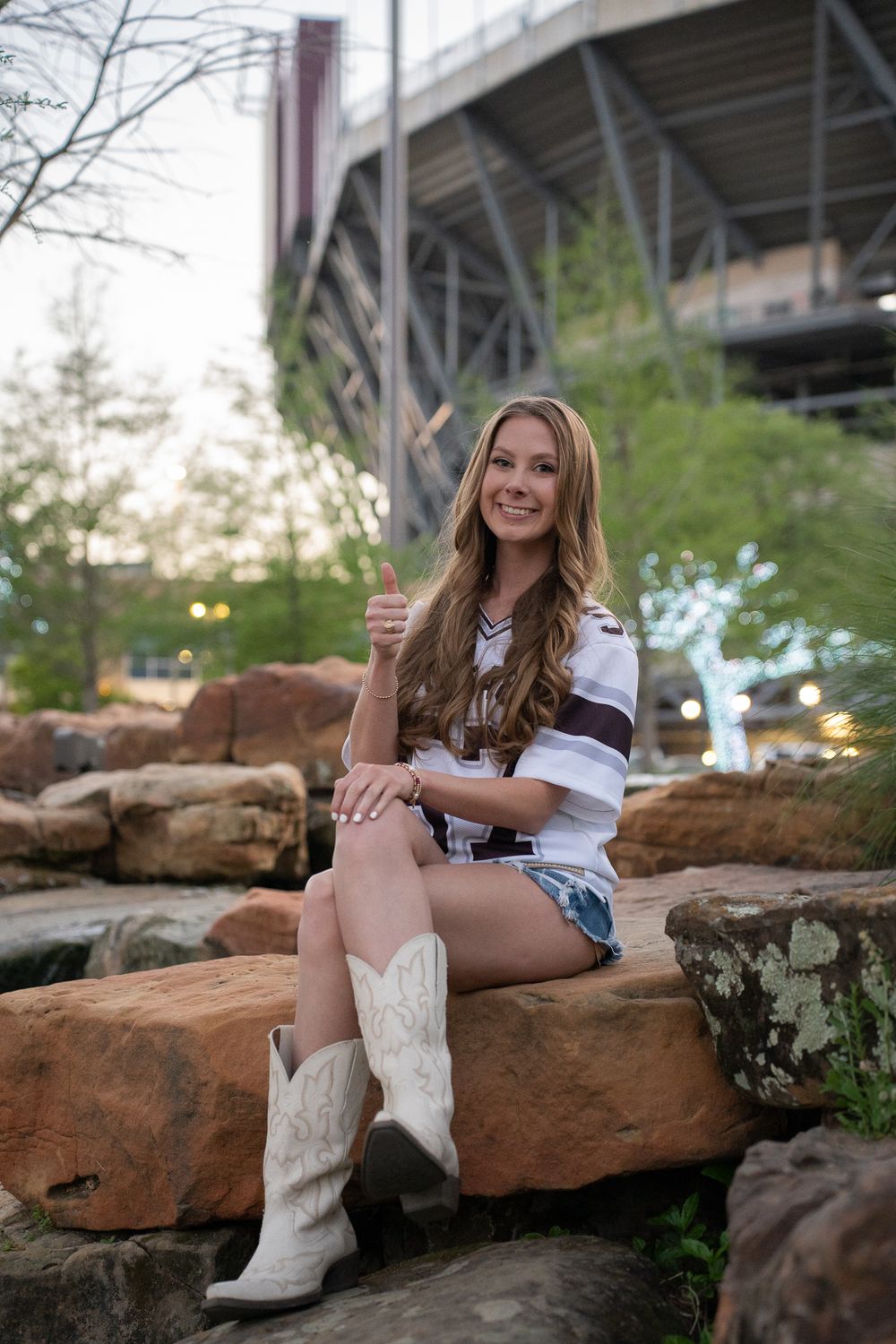 Person sitting on red rocks wearing a striped shirt and white cowboy boots outside a stadium.