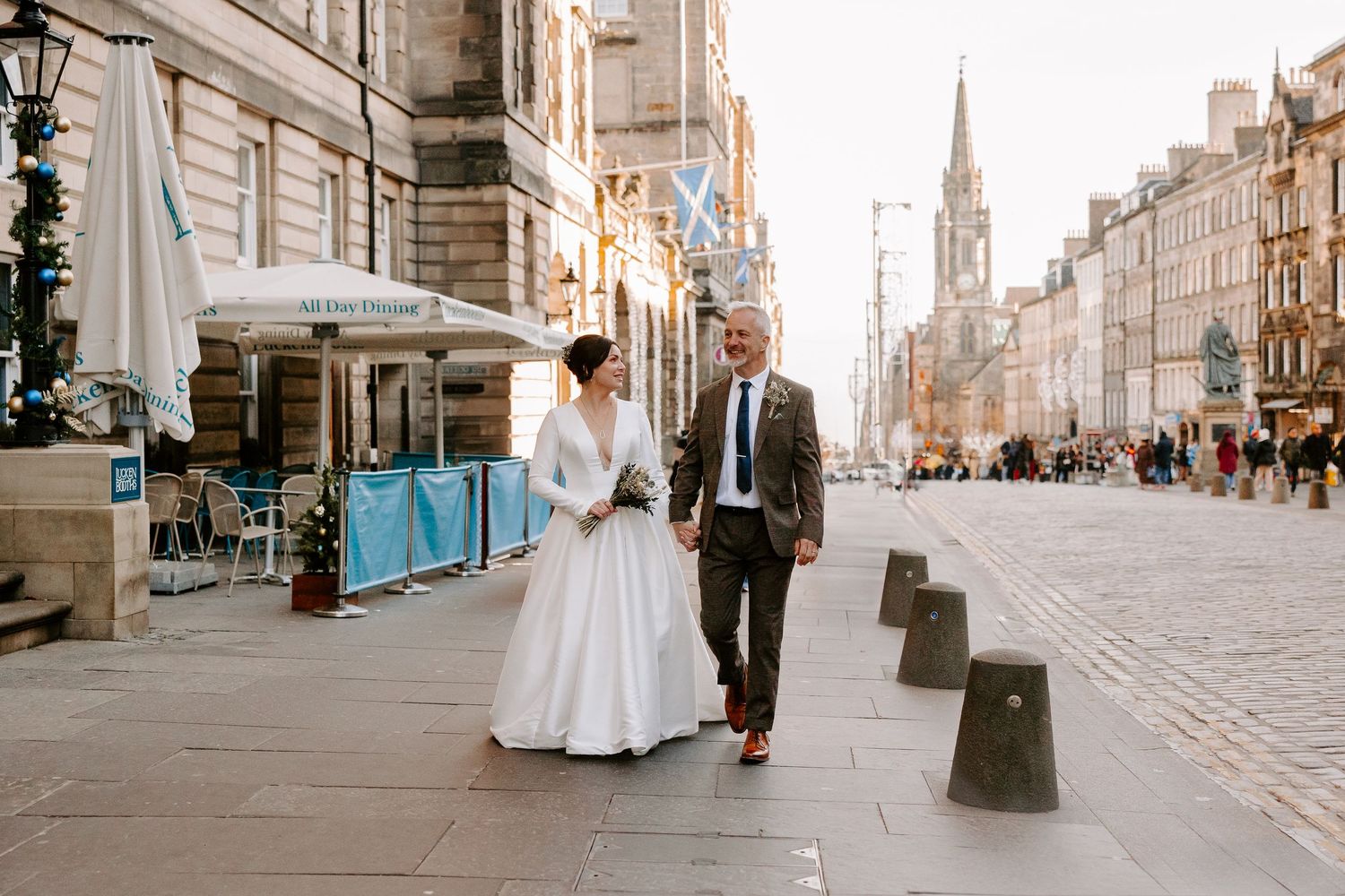 A wedding couple walks along Edinburgh's Royal Mile with the historic St Giles Cathedral spire visible in the background.