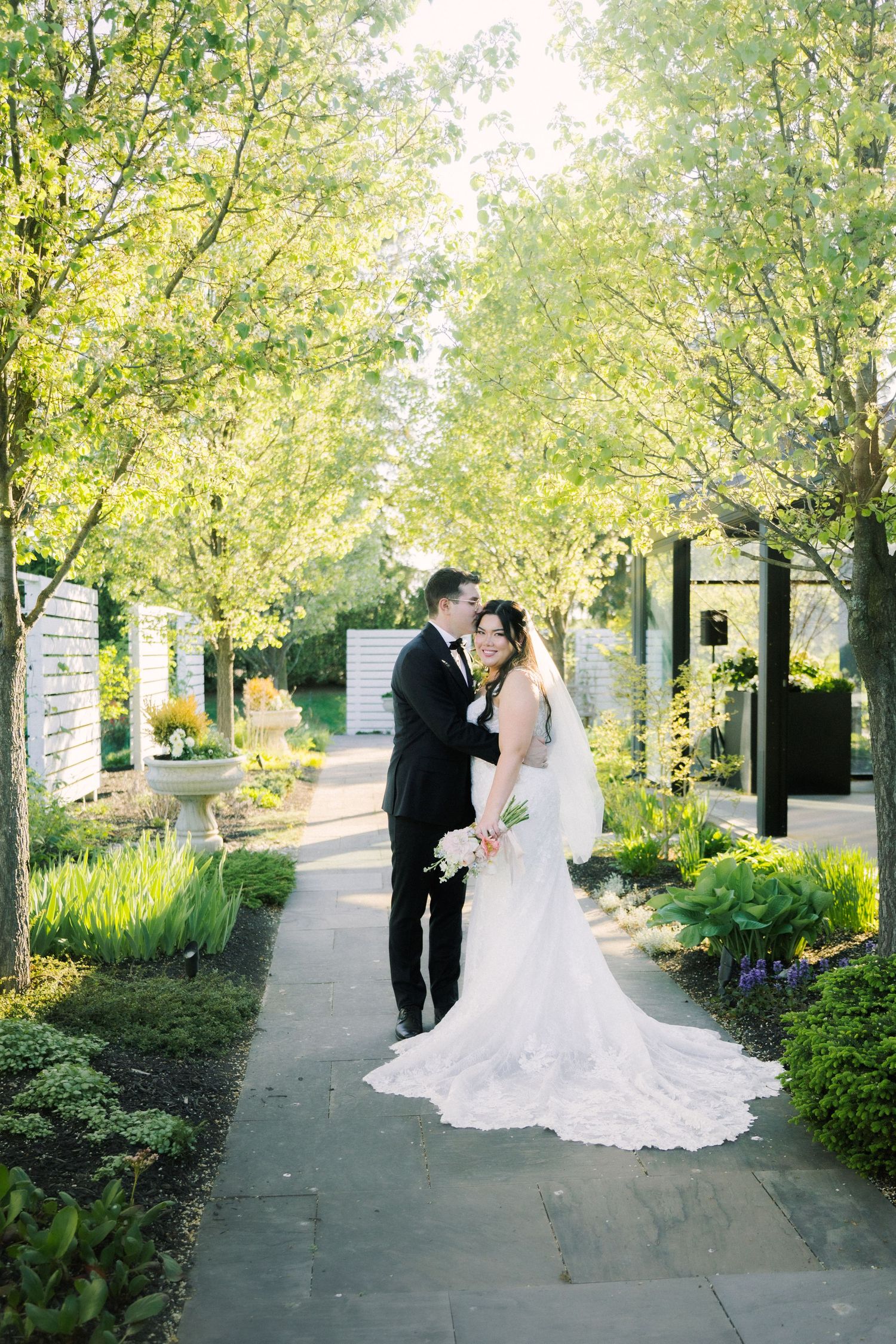 A couple poses in their wedding attire on a garden path lined with lush greenery and spring blooms.