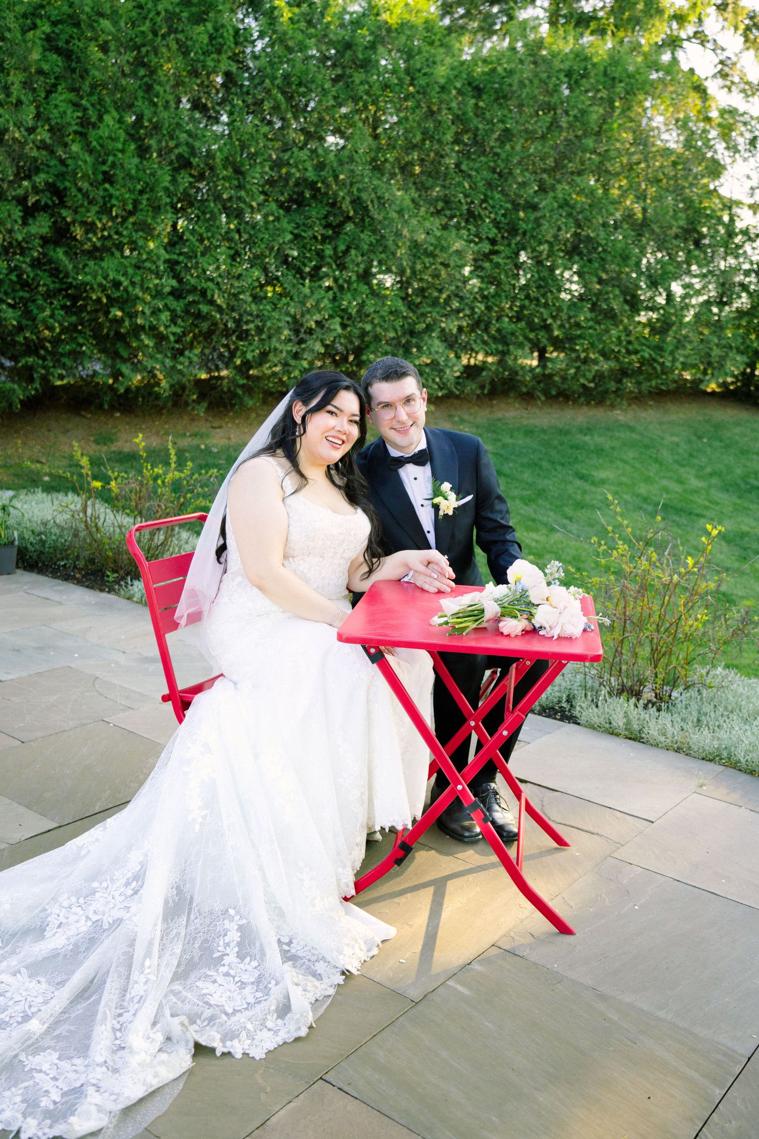 Bride and groom seated at red bistro table in garden setting with flowing white lace dress.